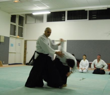 Un pratiquant en hakama accompagne son partenaire vers le sol ; derrière eux, d’autres élèves regardent, assis.
